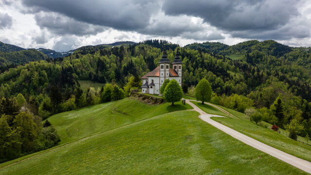 Aerial view of Cerkev svetega Volbenka nad Logom on a lush green hill surrounded by dense forests and rolling landscapes in Skofja Loka, Skofja Loka, Slovenia.