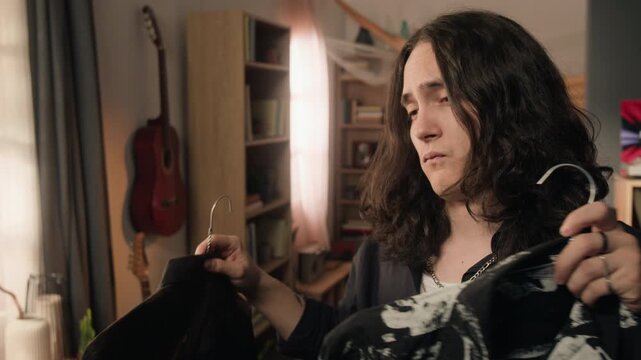 Tilt up shot of young man holding two patterned shirts on hangers, comparing them and looking undecided while choosing outfit. Perfect for fashion retail, wardrobe styling, or lifestyle vlogs