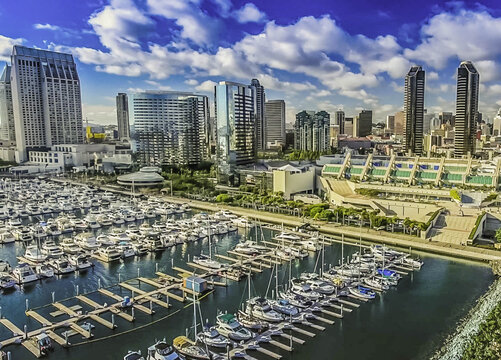Aerial view of the San Diego Marina with numerous yachts and boats docked at the piers, set against the modern city skyline under a bright blue sky. San Diego, California, United States.