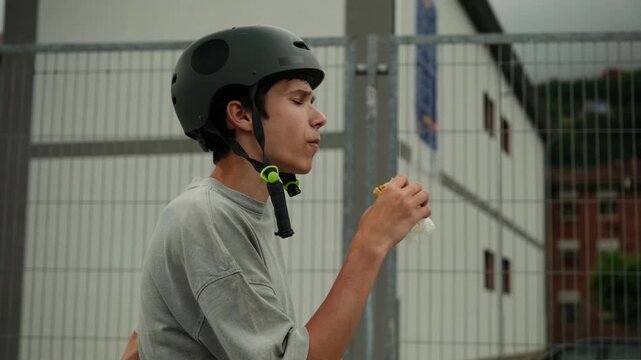 A teenage boy wearing a helmet takes a break to eat a sandwich at an outdoor skatepark.