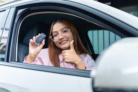 Happy Asian woman driver showing a smart key and giving thumbs up inside her electric car. Concept of new EV ownership finance approval and auto insurance.