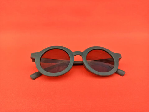 A pair of dark framed round sunglasses featuring red tinted lenses is centered on a bright, solid orange background. Minimalist, modern accessory for summer or fashion concepts.