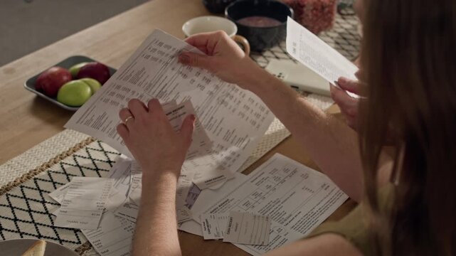 Over shoulder shot of Caucasian young adult couple reviewing utility bills and sorting receipts at table, pointing at financial document. For personal finance, budgeting apps, and debt management ads