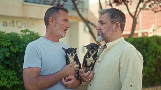 Two men hold chihuahuas, touch lips in a brief kiss and smile while facing each other in front of a residential building; affection companionship.