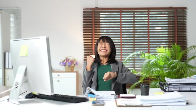 Asian businesswoman looking at her watch and celebrating the end of the working day with excitement and happiness while sitting at her messy desk in a modern office