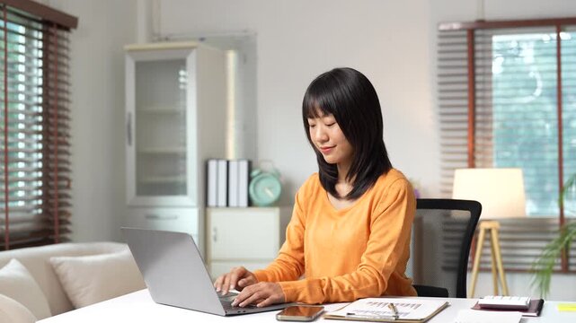 Young smiling asian businesswoman typing on laptop computer working on financial report at home office, happy female freelancer responding to client email in living room