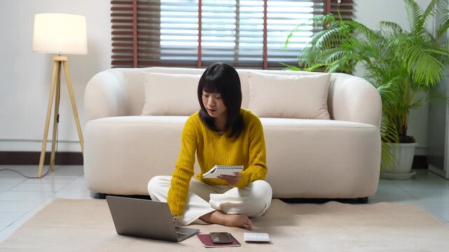 Focused asian woman studying online, taking notes and using her laptop for e-learning while sitting on the floor of her modern living room at home