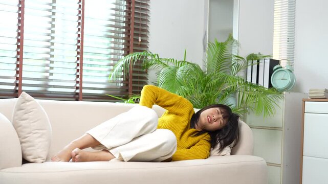 Asian woman suffering from acute lower back pain while lying on the sofa, experiencing muscle spasms, sciatica, and severe discomfort in her modern living room at home