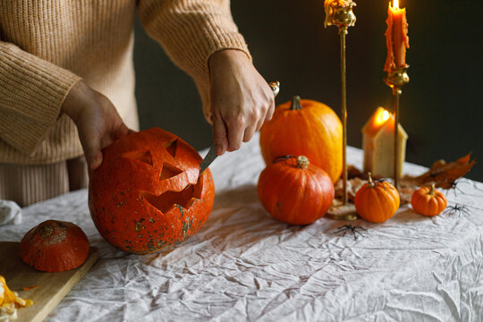 Carving halloween pumpkin. Woman hands holding pumpkin and knife on table with burning candles and spiders on moody background. Making jack o lantern, halloween preparation and decorations