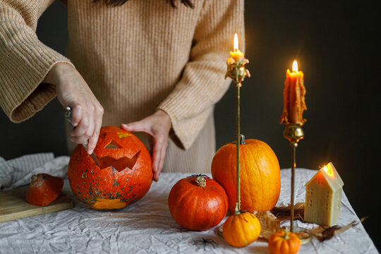 Happy Halloween. Woman carving halloween pumpkin on table with scary decorations and burning candles on dark moody background. Space for text. Making jack o lantern