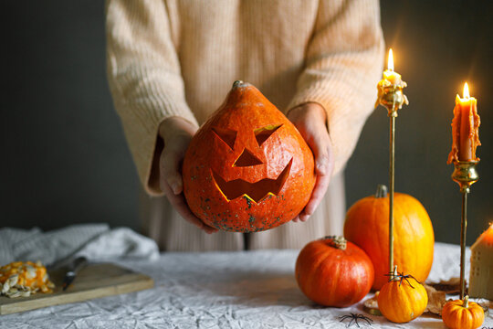 Happy Halloween. Woman holding carved halloween pumpkin on table with scary decorations and burning candles on dark moody background. Space for text. Making jack o lantern