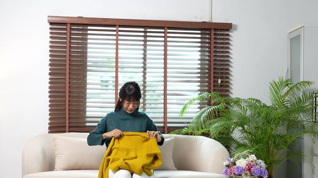 Diligent asian woman folding a yellow sweater while sitting on a sofa in a bright living room, showcasing a meticulous housekeeping and decluttering routine at home