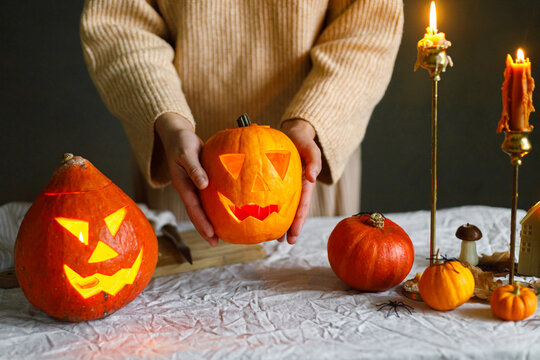 Happy Halloween. Woman carving halloween pumpkin on table with scary decorations and burning candles on dark moody background. Space for text. Making jack o lantern