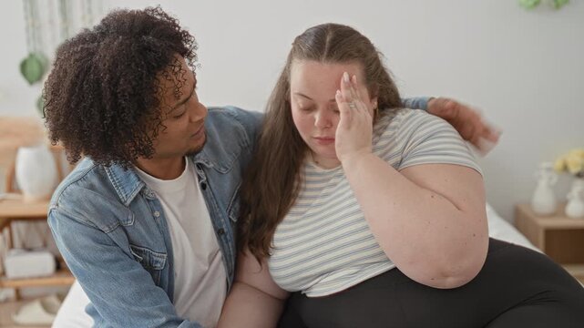 Woman holds hand to forehead on bed while man wraps an arm around her shoulder in a bedroom, leaning close in a consoling embrace; comfort support intimacy.