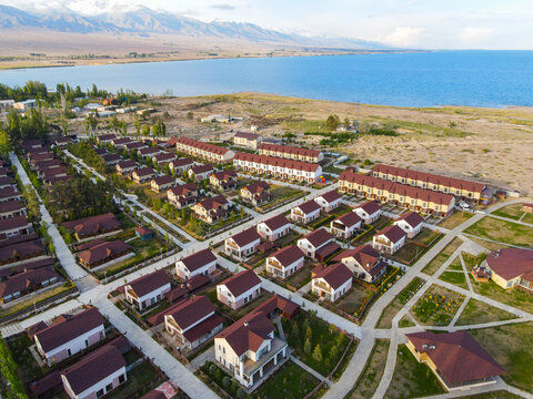 Beautiful aerial view of coastal cottages and houses with red roofs located on the sandy shore of Issyk Kul Lake with distant mountains under a blue sky