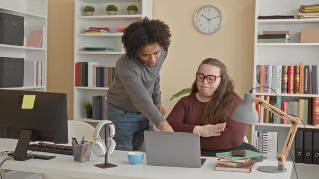 Man points to laptop and shows papers while seated woman watches with folded arms in an office workspace with bookshelves and a clock; mentoring focus.