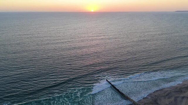 A dramatic sunset aerial shot capturing the international border wall stretching from the coastal landscape directly into the breaking waves of the Pacific Ocean.
