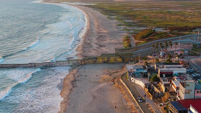 A striking high-angle aerial view of the industrial steel international border fence extending directly into the breaking waves of the Pacific Ocean.