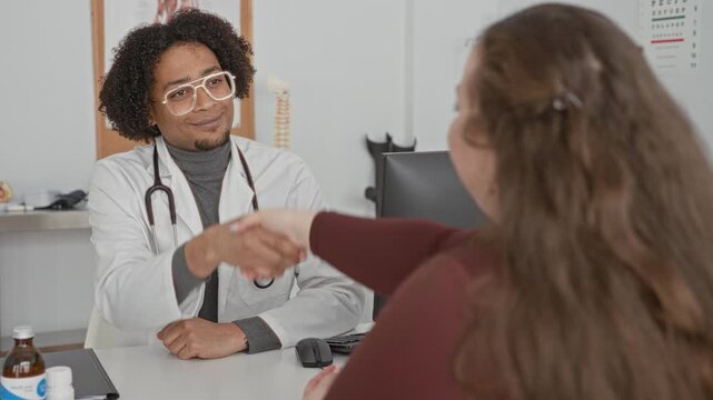 Man doctor in white coat with stethoscope at desk shaking patient's hand, hands clasped while speaking with woman patient in clinic, monitor and spine model visible; reassurance care.