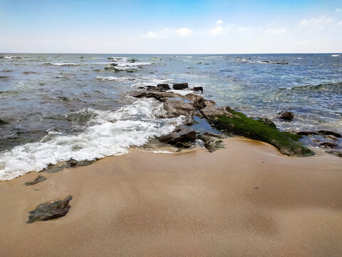 Experience serene solitude on a peaceful, empty beach where gentle waves crash over mossy rocks under a blue sky. Evoking tranquility and natural beauty, with nobody in sight.