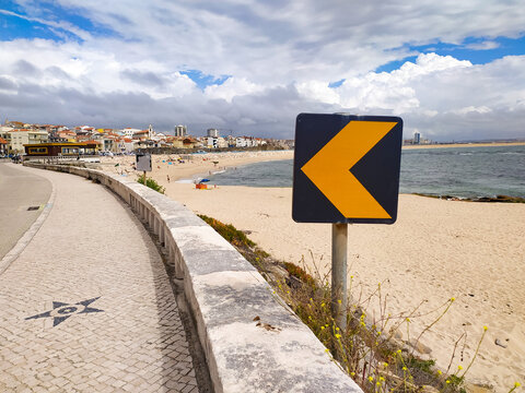 A vivid yellow arrow sign guides the way along a scenic coastal promenade overlooking a quiet sandy beach and blue ocean. Nobody in sight, evoking a peaceful sense of wanderlust.