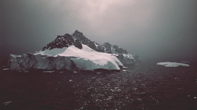 Secluded ice island surrounded by dark water, brooding low clouds above jagged peaks, scattered ice fragments add foreground interest, scene feels cinematic,