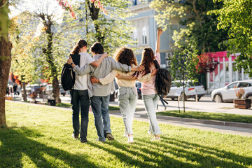 Obraz premium Group of four young adults walking together in a park, arms around each other, with green grass and trees, city buildings visible in the background