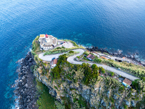 Aerial view of Farol do Arnel perched on a rugged cliffside with a winding road, lush vegetation, and the deep blue Atlantic Ocean in Nordeste, Acores, Portugal.