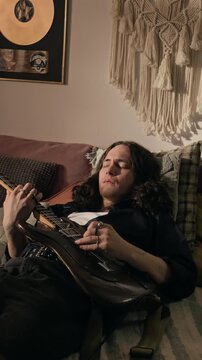 Vertical video of young man lying on sofa and playing electric guitar. Footage for music streaming ads, online lessons, or creative lifestyle marketing