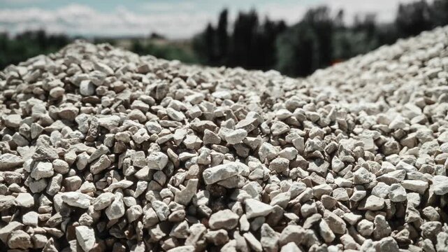 Sunlit low-angle view of crushed stone stockpiles, gravel piles, and rough aggregate mounds at outdoor concrete production site with industrial textures, depth, and open daylight