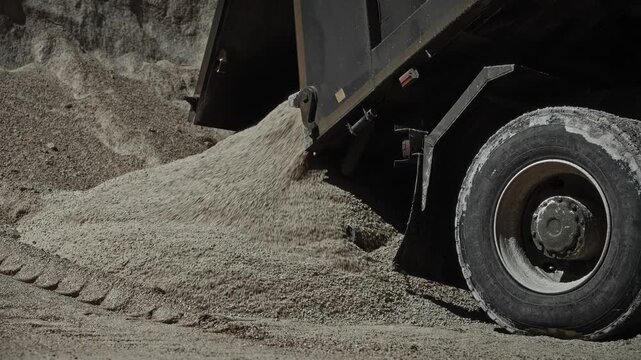 Heavy dump truck unloading sand into growing mound at outdoor aggregate yard, bright daylight, close view showing flowing material, rugged vehicle, industrial worksite detail