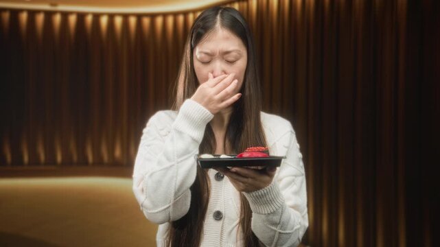 Young chinese woman holds sushi plate and pinches nose at a piece of sushi in studio; disgust aversion hesitation.