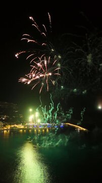 Vertical aerial view of a fireworks display over the port of Javea on the Costa Blanca in Alicante, Spain. Green firework bursts with trailing willow-effect sparks light up the night sky above an illu
