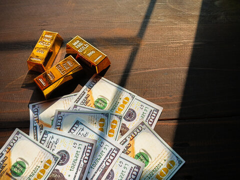 Gold bars and US dollar banknotes on wooden table with sunlight, symbolizing wealth, investment, finance, savings, and safe assets.