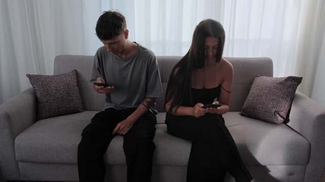 Social media replacing real connection. Young couple sitting together on sofa while focused on smartphones, online engagement, emotional disconnection and reduced face to face communication