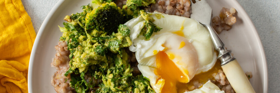 A healthy breakfast bowl features green buckwheat topped with pesto, steamed broccoli, and poached eggs. A fork rests beside the dish ready for a nutritious meal, banner