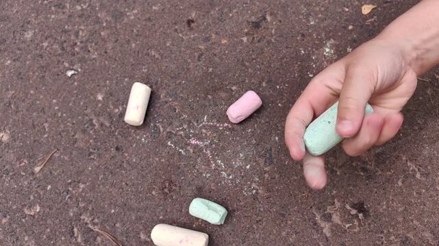 Child chooses a green sidewalk chalk, crushes it between small hands and lets colorful chalk dust fall onto asphalt, creative outdoor play, summer childhood, artistic exploration
