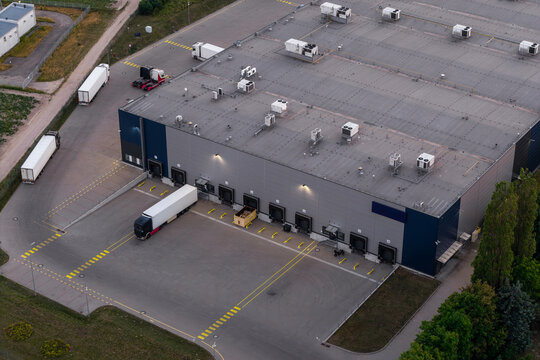 Aerial view of a large distribution center warehouse featuring multiple loading docks, parked semi-trucks, and a paved logistics yard in Warszawa, Mazowieckie, Poland.