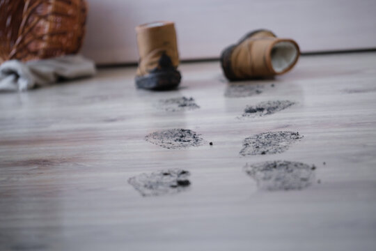 Footprints lead across a wooden floor next to old boots and a rolled blanket on the gray floor. The scene shows a relaxed indoor space with personal items scattered around.
