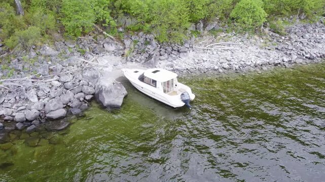 White cabin motorboat moored against rocky lakeshore, outboard engine visible, shallow clear water, overcast light, high-angle view showing stones, shoreline, driftwood, forest edge