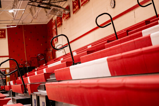 Rows of empty red tiered bleacher seating with black safety handrails extend across a school gymnasium, photographed at an angle. No people, capturing anticipation before a game or assembly.