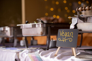 The word "vegan" written on a chalkboard sign on a wooden easel in front of a row of stainless steel chafing dishes. No people. © Jeanne Sager Photo
