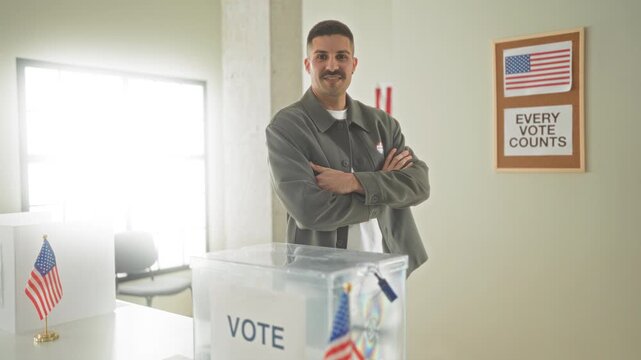 Man with arms crossed beside a transparent ballot box, small american desk flag and every vote counts sign on the wall in a building; patriotic pride.