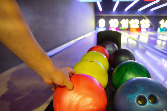 A close-up view of a hand selecting a red bowling ball from a rack in a modern bowling alley with colorful balls and neon-lit lanes in the background.