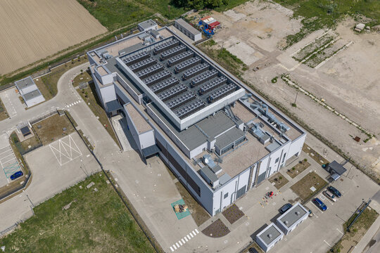 Aerial view of a modern data center facility with cooling units on the roof, surrounded by parking areas and green fields in Warszawa, Mazowieckie Voivodeship, Poland.
