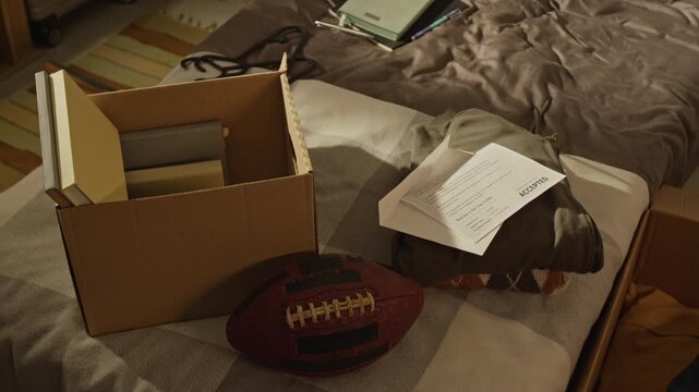 No people shot of cardboard box packed with books, football, and college acceptance letter sitting on bed, showing student preparation for moving. Perfect for university marketing, student loan ads