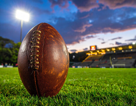 A leather Gaelic football sits on the grass of a community pitch under the bright lights of a stadium evening.