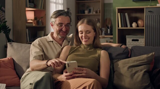 Medium shot of middle aged Caucasian couple sitting on sofa, browsing smartphone and pointing at screen. Footage suitable for mobile application promos or online banking advertisements
