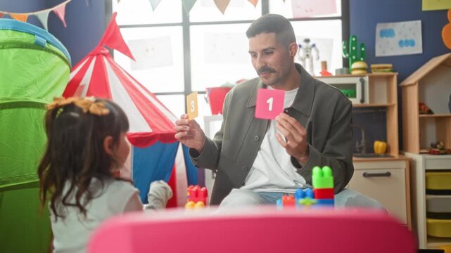 Teacher holds number card while child points finger to card in building learning space; learning joy.