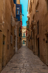 Obraz premium Narrow Alleyway road in valletta, malta with limestone buildings lining each side with colorful balconies and windows and clear blue summer skies overhead